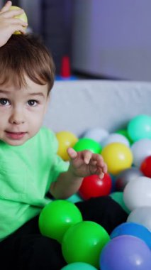 Güzel çocuk kuru havuzda topların arasında oturur. Neşeli, enerjik bir çocuk, yan tarafa top atıyor. Arkaplan bulanık. Dikey video