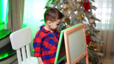 Kareli tişörtlü çocuk tahtaya resim çiziyor. Güzel Noel ağacı arka planda.