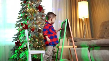 Sevimli çocuk Noel ağacının yanındaki tahtada duruyor. Bebek tahtaya tebeşirle resim çiziyor..
