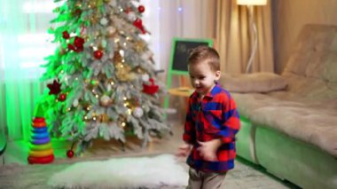 Dört yaşında, kareli tişörtlü sevimli bir çocuk odada dans ediyor. Bebeğim Noel ağacının yanında eğleniyor..