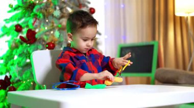 Kırmızı ve mavi tişört giyen sevimli erkek bebek masada oturuyor. Yaratıcı çocuk hamuru makasla keser. Arkaplanda Noel ağacı.