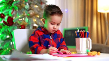 Kareli tişörtlü sevimli çocuk keçeli kalemle resim çiziyor. Bebek evde sanat yapıyor. Arkaplanda Noel ağacı.