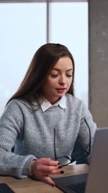 Genç, uzun saçlı kadın bilgisayara odaklanmış görünüyor. Elde bardak tutan ofis çalışanı dizüstü bilgisayarla çalışır. Dikey video