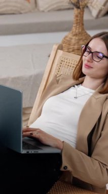 Koyu renk saçlı kadın koltuğa rahatça uzanıp oturur. Bayan rahat atmosferde dizüstü bilgisayarla çalışıyor. Dikey video