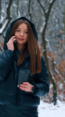 Bir fincan sıcak kahveyle kış parkında yürüyüş. Koyu renk saçlı genç bayan dışarıda dolaşırken telefonda konuşuyor. Dikey video