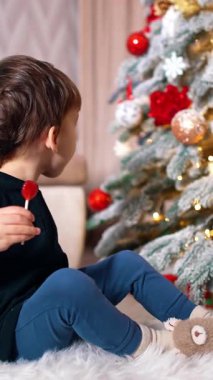 Lolipopu olan sevimli erkek bebek Noel ağacında oturuyor. Bebek ağaçtaki güzel süslemelere dokunuyor. Dikey video