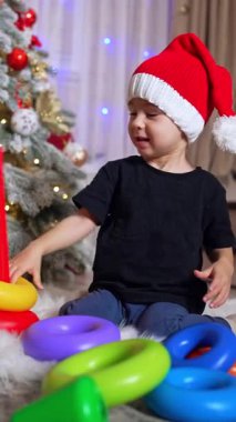 Kırmızı şapkalı yakışıklı çocuk piramitle oynuyor. Çocuk güzel Noel ağacının yanında oyuncakla oynuyor. Dikey video