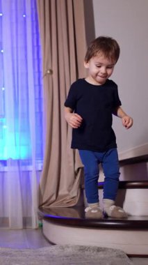 Evdeki merdivenlerden atlayan sağlıklı küçük bir çocuk. Evde güzel bir Noel ağacıyla süslenmiş güzel bir bebek. Düşük açı. Dikey video