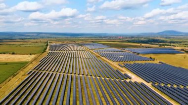 Yeşil bir vadideki güneş enerjisi panelleri. Bulutlu gökyüzünün altındaki tepelerle çevrili yeşil bir vadi boyunca uzanan güneş panellerinden oluşan hava aracı görüntüleri..