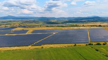 Güneş çiftliklerine ve açık kırlara geniş bir bakış açısı. Etrafı kırsal ve uzak tepelerle çevrili geniş bir güneş enerjisi tesisinin panoramik hava görüntüsü.