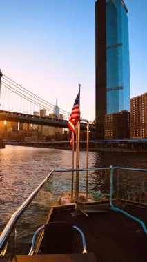 Teknenin önünde Amerikan bayrağı var. New York 'un Skyline' ı ve Manhattan Köprüsü 'nün arka planında göz kamaştırıcı batan güneş. Dikey video.