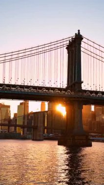 Binalar ve Manhattan Köprüsü arasında güneş batıyor. Tren köprüden geçiyor. Düşük açı. New York, ABD. Dikey video.