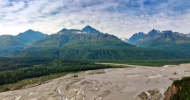 Alaska, ABD 'de vahşi doğa üzerinde uçuş. Sığ nehir deltası, ormanlık vadi ve yemyeşil dağlar. Gökyüzü bulutlu.