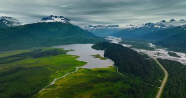 Güzel yeşil vadideki nehre yaklaşıyoruz. Arkaplanda bulutlarla kaplı karla kaplı dağlar. Alaska, ABD 'de kasvetli bir gün. Hava görünümü.