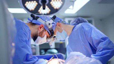 Cerrahi lensler takan kendine güvenen erkek cerrahlar ameliyat yapıyor. Beyin cerrahları modern aletler kullanır. Arka planda yuvarlak lamba.