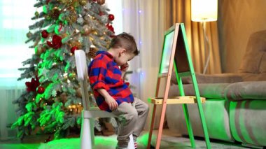 Noel ağacının yanında oturan çocuk düşünüyor. Kırmızı ve mavi tişörtlü küçük çocuk Noel ağacının yanında otururken düşünceli görünüyor..