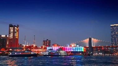 Gemi geceleri Doğu Nehri 'nin sularından geçiyor. 17. rıhtımdaki barın gökkuşağı aydınlatması ve arka plandaki Brooklyn Köprüsü..