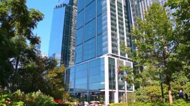 New York, ABD, 5 Ağustos 2025: Dev, yüksek binaların yakınında yeşil ağaçlar yetişiyor. Gökdelenin cam cepheleri mavi gökyüzünü yansıtır. Düşük açı görünümü.