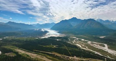 Parlak güneş tarafından göz kamaştıran dağların çarpıcı manzarası. Beyaz bulutlar gökyüzünü kaplıyor. Dallanan nehir vadide akar. Hava görüntüsü. Alaska, ABD.
