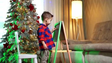 Kareli tişörtlü çocuk tahtaya yazı yazıyor. Güzel Noel ağacı arka planda.