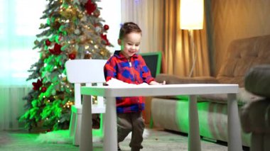 Sevimli neşeli çocuk masayı sandalyeye çekti ve oturdu. Arkaplanda süslenmiş Noel ağacı.