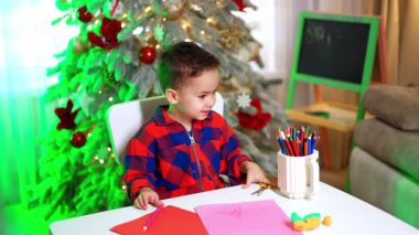 Çekici çocuk Noel ağacının yanındaki masada oturuyor. Küçük çocuk makası ve pembe keçeli kalemi alır..