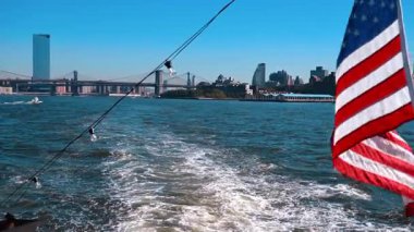 Nehir kıyısında hareket eden teknede bayrak ve ampuller var. Brooklyn Köprüsü 'nün, Manhattan Köprüsü' nün ve şehrin ufuk çizgisinin görüntüsü.
