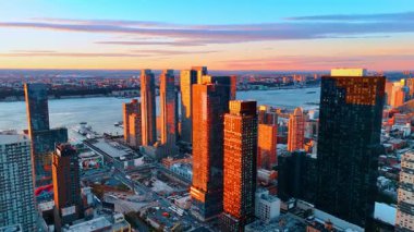 Parlak turuncu ışık günbatımında gökdelenlerin cephelerini aydınlatıyor. İHA görüntülerinden New York mimarisi nehir kıyısında..