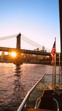 Önünde Amerikan bayrağı olan tekneden bir görüntü. Arkaplanda Manhattan Köprüsü ve ufuk çizgisi arasında güneş batıyor. Dikey video.