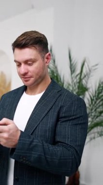 Koyu saçlı, beyaz ceketli erkek akıllı telefonuna bakıyor. Uzaktan kumandayla çalışan kendinden emin bir girişimcinin portresi. Dikey video