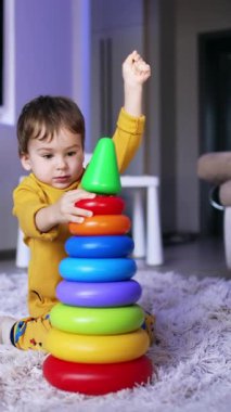 Sarı pijamalı şirin bebek oyuncak piramidini sökmeye çalışıyor. Evinde oynayan sevimli çocuk. Dikey video