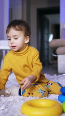 Yumuşak ekose üzerinde oturan küçük beyaz çocuk bir oyuncakla oynuyor. Küçük çocuk piramit topluyor. Dikey video