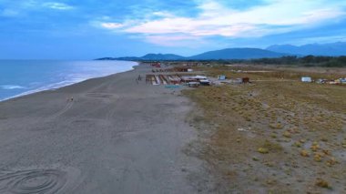 Adriyatik Denizi kıyısındaki kumlu plajın geniş hattı. Güneş yatakları ve barlarla turist altyapısına yaklaşıyoruz. Arkaplanda sıradağlar var. Karadağ. Hava görünümü.