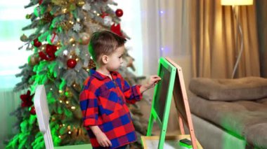 Sevimli küçük çocuk tahtada tebeşirle yazı yazıyor. Arkaplanda süslemeleri olan güzel Noel ağacı.