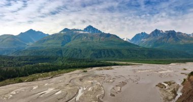 Ormanlık vadide akan sığ nehir deltasından aşağı iniyoruz. Arka plandaki tepeli yemyeşil dağlar. Gökyüzü bulutlu. Alaska, ABD.