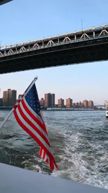 Boat with an American flag moving by the river. Low angle view at the Manhattan Bridge with a train moving by. Vertical video.