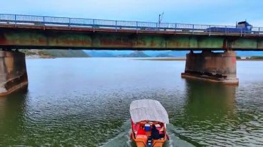 Ulcinj, Montenegro, 14 August 2025: Boat Approaching Old Bridge. A red boat moves toward an old rustic bridge across a quiet lake.