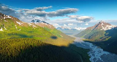 Sunlit Alaska valley with braided river. Golden light casts shadows over dense forest and braided river beneath snowy peaks.