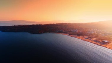 Suyun üzerinden uçarak yerleşim olan kıyıya yaklaşıyoruz. Yumuşak turuncu ışık gökyüzünü ve ağaçlık sahili boyuyor..