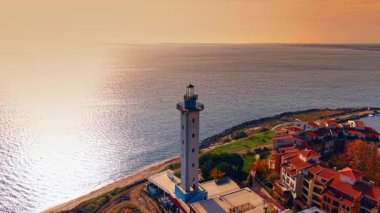 Residential neighborhood of colorful houses and lighthouse located at the sea coast. Bright sun dazzles the waterscape at backdrop. Aerial perspective.