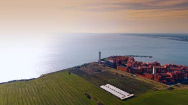 Footage over the green field at the coast of the sea. Approaching colorful residential area with a lighthouse and pier.