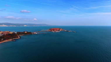 Approaching highly inhabited island with red-roofed houses connected by the dam with the mainland. Coastal part of Nessebar, Bulgaria surrounded by the Black Sea. Aerial view.