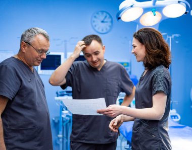 Cerrahlar hasta verilerini tartışıyor. Cerrahi odasındaki sağlık uzmanları çizelgeyi inceliyor.