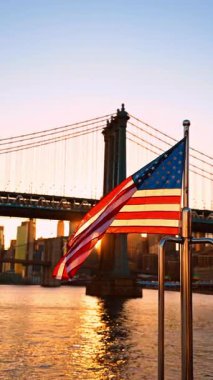 American flag gently waving at the wind. Setting sun among the buildings and the Manhattan Bridge at backdrop. Vertical video.