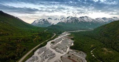 Footage above the shallow river flowing along the verdant mountains. Snow-covered rocks at backdrop under the cloudy sky. Alaska, USA.