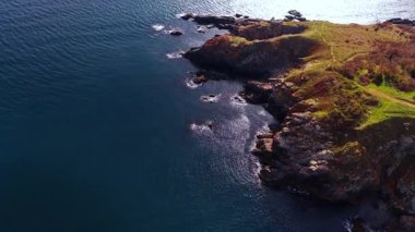 Rocky headland above deep blue sea. Steep coastal rocks meet dark shimmering water along a rugged grassy point.