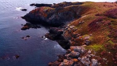 Sunlit cliffs rising from dark waters. Warm light highlights rugged rock faces meeting calm deep blue sea along the shore.