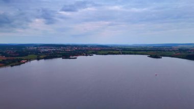 A wide expanse of lake water against the backdrop of distant evening city lights. A calm scene of a large lake with a sailboat at sunset under a gray, overcast sky.