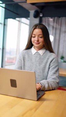 Focused Caucasian woman with long brown hair sits at the desk. Office employee types on her laptop and smiles. Vertical video