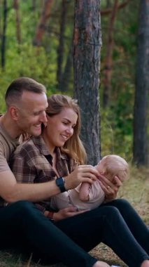Caucasian parents are happy to spend time together with their newborn baby. Couple sits in the forest holding a baby and smiling to it. Vertical video